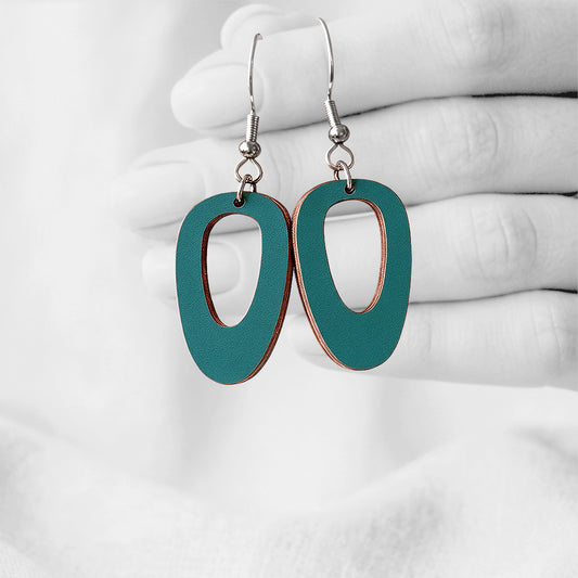 Petrol Luoto (Islet) plywood earrings made in Finland, photographed hanging from a hand in black and white against a softly blurred light background.