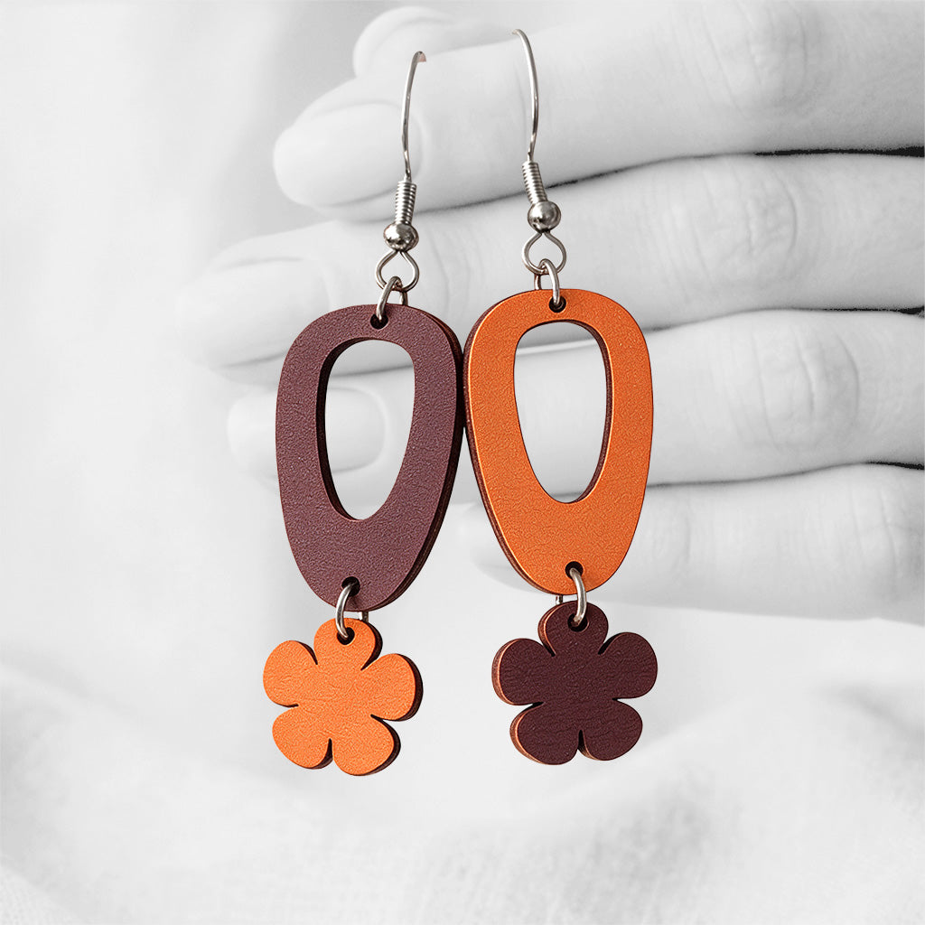 Orange and brown Luototar (Nature Goddess) plywood earrings handmade in Finland, photographed hanging against a black-and-white hand background.