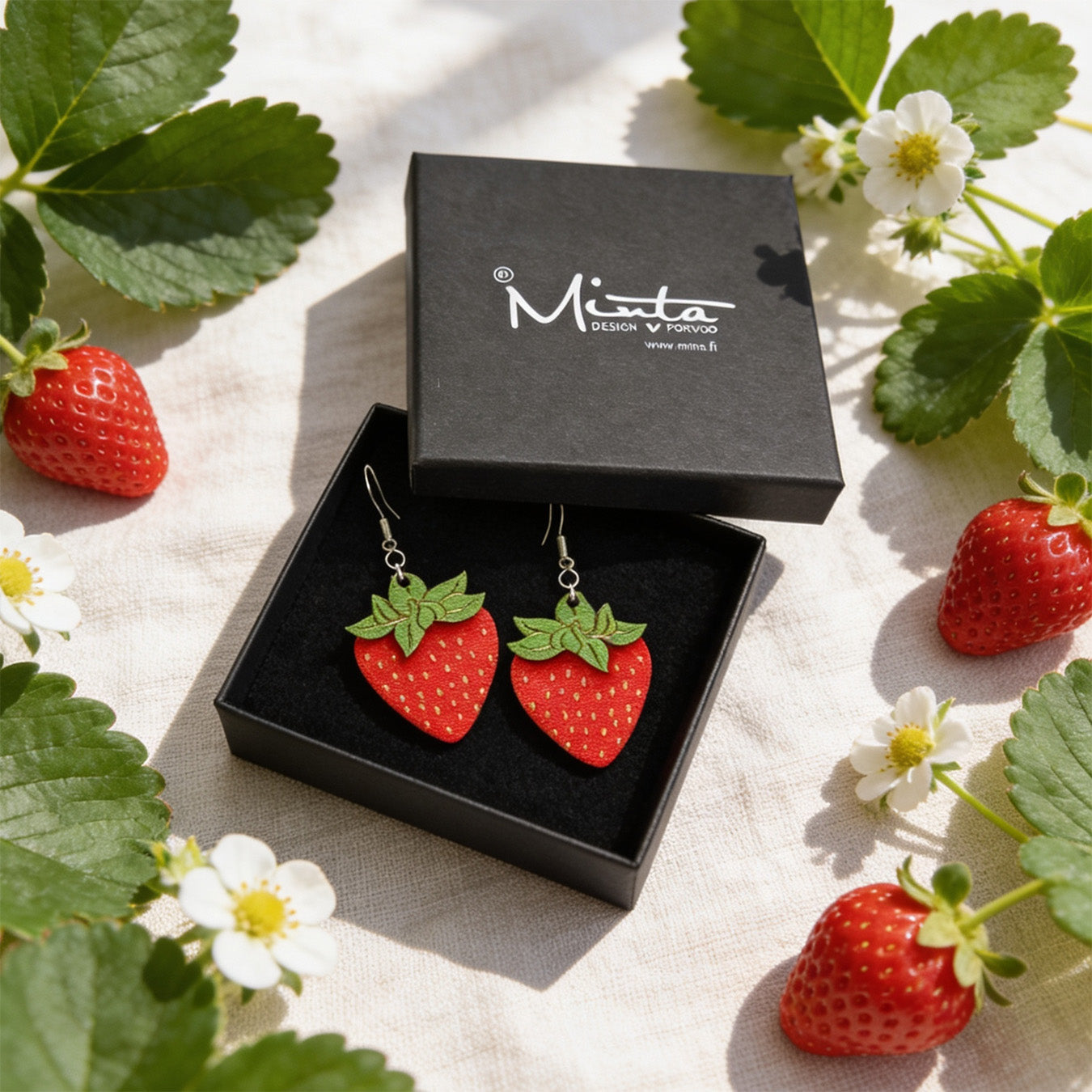 Handmade strawberry earrings displayed in a black jewelry box surrounded by fresh strawberry leaves, white strawberry flowers and ripe strawberries. Lightweight Finnish birch plywood berry earrings styled in bright natural daylight with a fresh Nordic summer atmosphere.