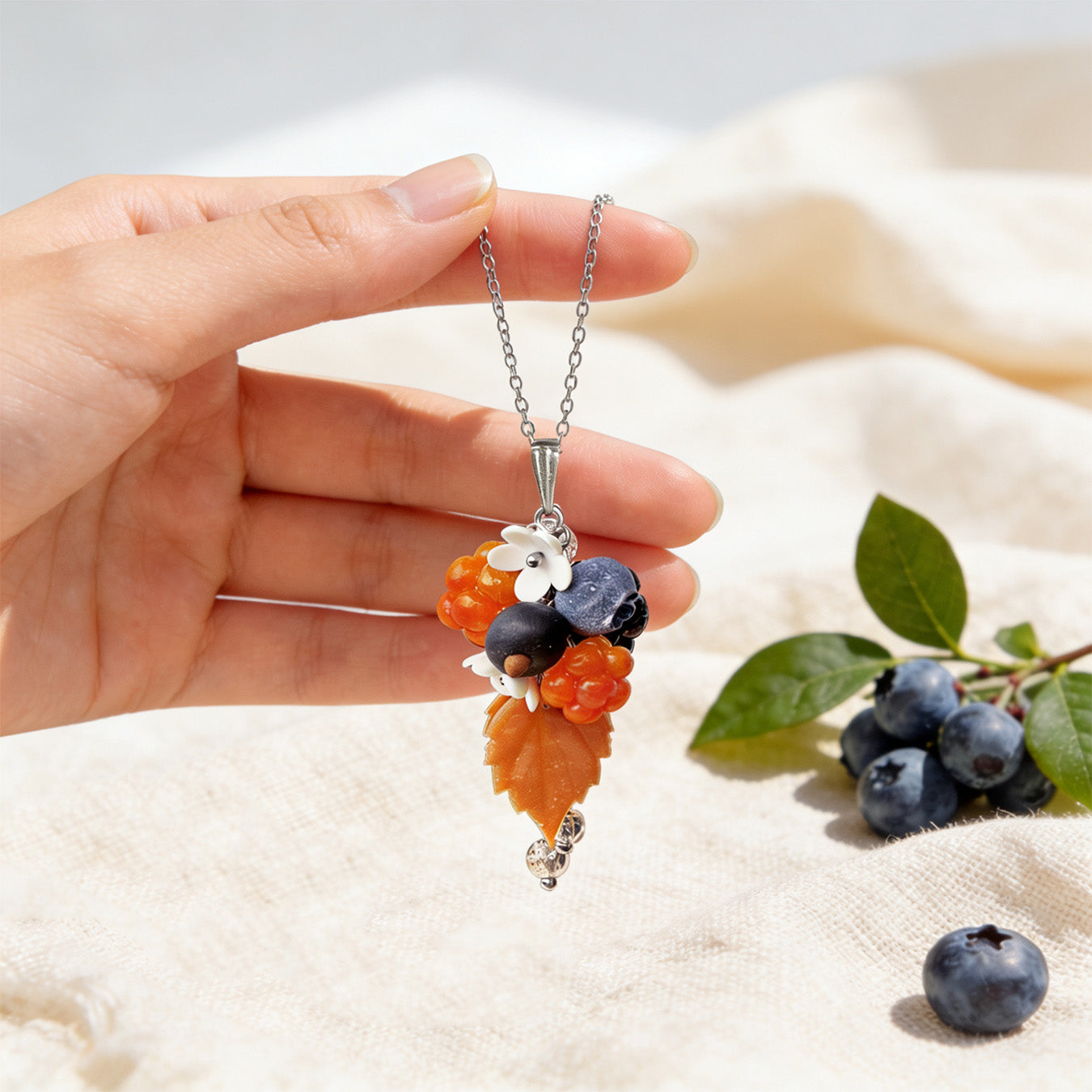 Berry Forest Necklace held gently in hand for scale, featuring hand-shaped berries and leaves made from polymer clay. Natural linen fabric, fresh blueberries, and green leaves in soft natural light. Handmade in Finland by byMinta.