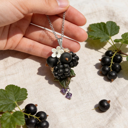 Hand holding a handmade berry necklace for size reference, featuring sculpted blackcurrants, blackberries, detailed green leaves, a small white flower and purple crystal accents. Surrounded by real black currants and currant leaves on soft linen in bright natural daylight.