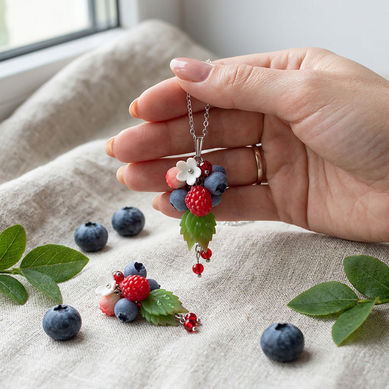 Berry Dance Necklace held gently in hand for scale, featuring hand-shaped berries and leaves made from polymer clay. Natural linen fabric, fresh raspberries, blueberries, and green leaves in soft natural light. Handmade in Finland by byMinta.