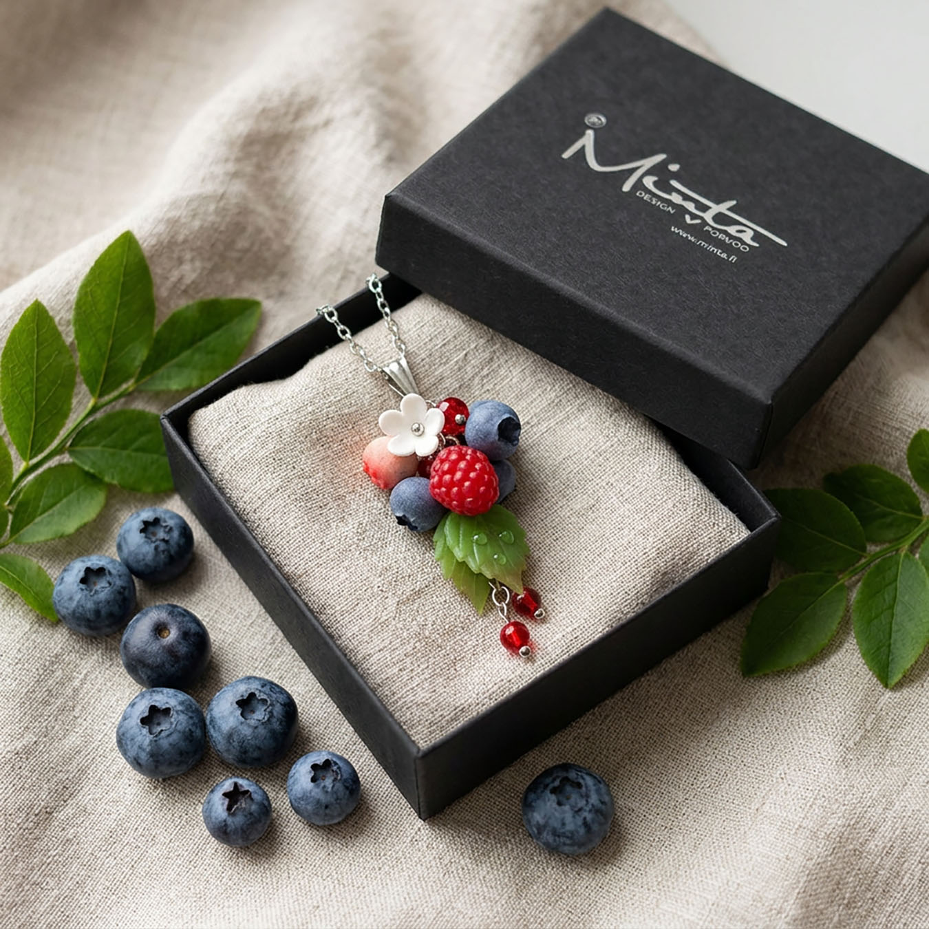 Berry Dance Necklace presented in a gift box, surrounded by natural linen fabric, fresh raspberries, blueberries, and green leaves in soft natural light. Handmade necklace with hand-shaped berries and leaves, handmade in Finland by byMinta.