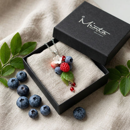 Berry Dance Necklace presented in a gift box, surrounded by natural linen fabric, fresh raspberries, blueberries, and green leaves in soft natural light. Handmade necklace with hand-shaped berries and leaves, handmade in Finland by byMinta.