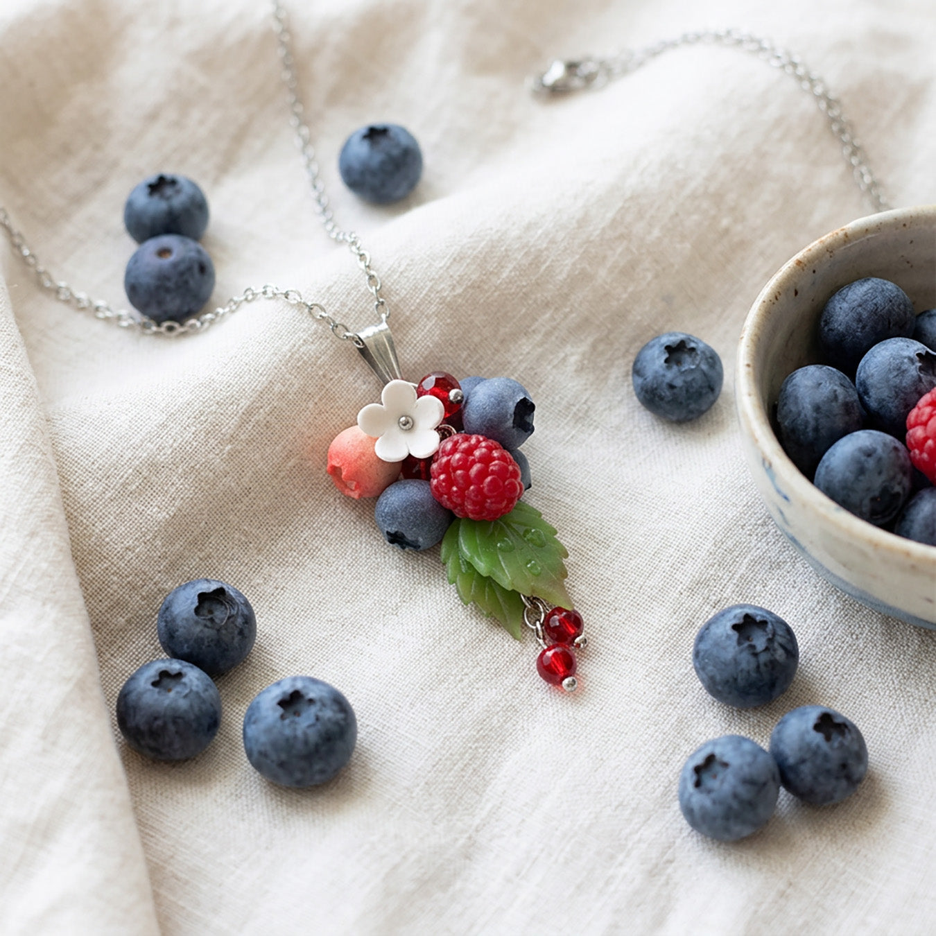 Berry necklace with handmade raspberry, blueberries and white flower on linen fabric, surrounded by fresh blueberries, handcrafted Finnish jewelry.