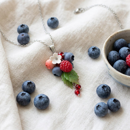 Berry necklace with handmade raspberry, blueberries and white flower on linen fabric, surrounded by fresh blueberries, handcrafted Finnish jewelry.