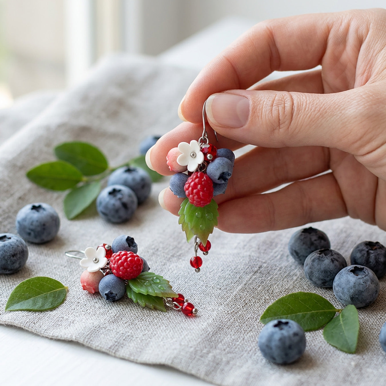 Dancing Berries Earrings photographed with fresh berries and green leaves in soft natural light, with one earring gently held by hand for scale. Handmade in Finland by byMinta.