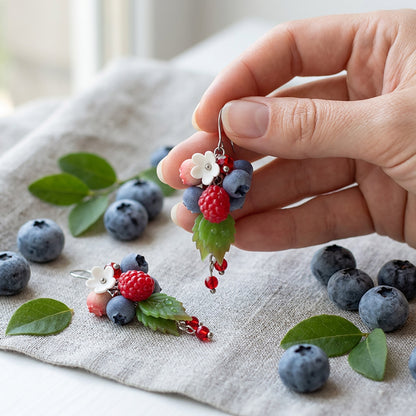 Dancing Berries Earrings photographed with fresh berries and green leaves in soft natural light, with one earring gently held by hand for scale. Handmade in Finland by byMinta.