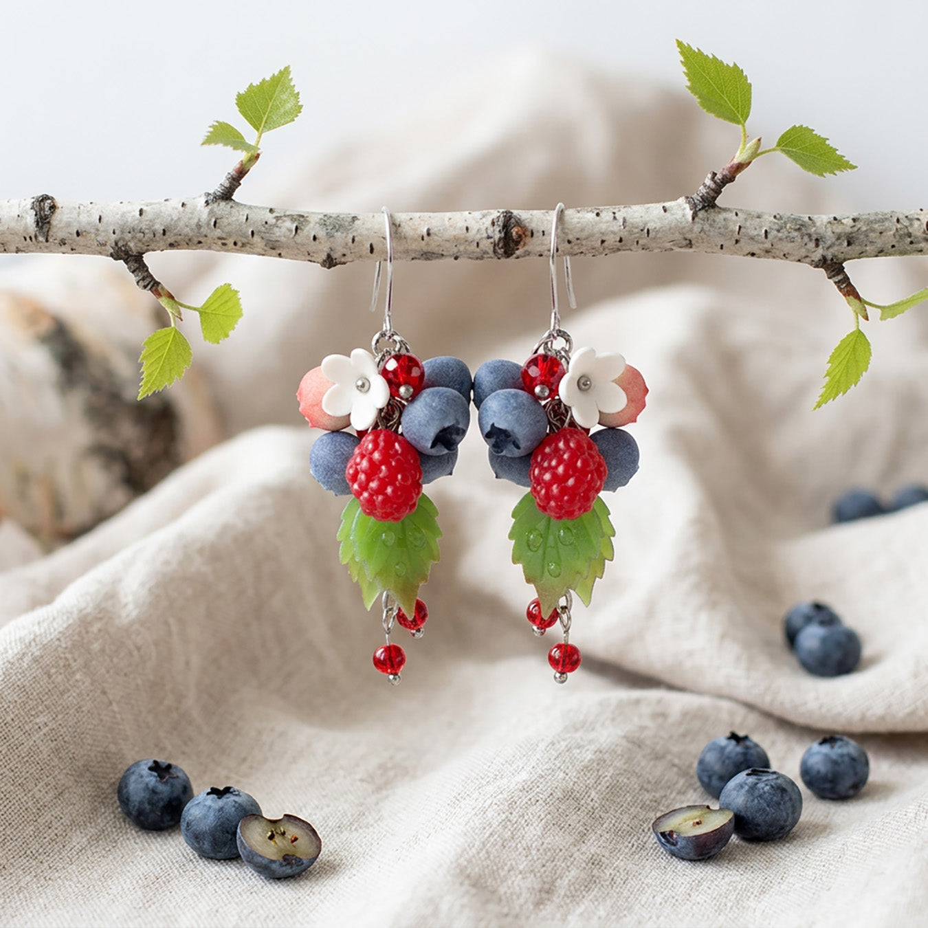 Berry earrings with handmade raspberries, blueberries and white flowers hanging from a birch branch, linen fabric and fresh berries in the background, handcrafted Finnish jewelry.