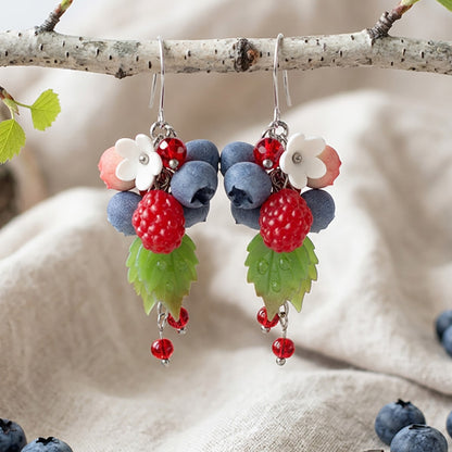 Handcrafted Marjatanssi earrings featuring raspberries, blueberries and flowers, hanging from a birch branch in soft natural light.
