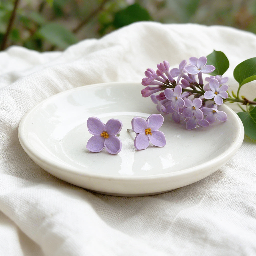 Lilac flower stud earrings made in Finland, displayed on a white ceramic plate with lilac blossoms and soft white fabric.