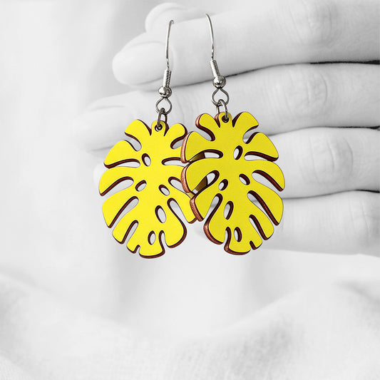 Yellow Monstera plywood earrings made in Finland, photographed hanging from a hand in black and white against a softly blurred light background.