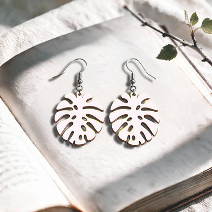 White Monstera plywood earrings handcrafted in Finland, photographed on a vintage book in natural sunlight