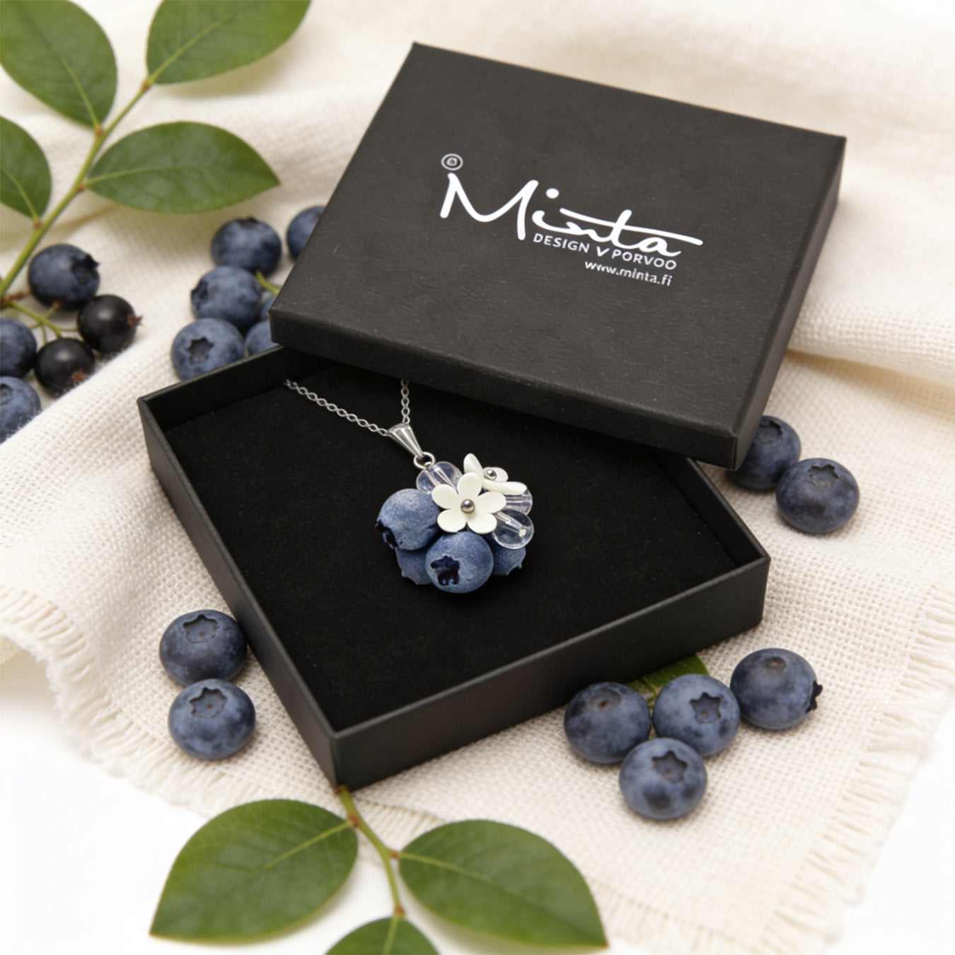 Blueberry Bouquet Necklace presented in a gift box, surrounded by natural linen fabric, fresh blueberries, and green leaves in soft natural light. Handmade necklace with blueberries and delicate flowers, handmade in Finland by byMinta.