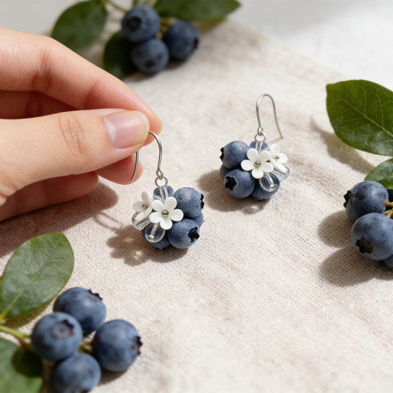 Blueberry Bouquet Earrings photographed with fresh blueberries and green leaves in soft natural light, with one earring gently held by hand for scale. Handmade in Finland by byMinta.