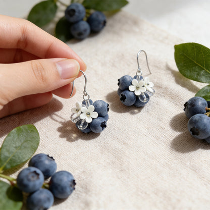 Blueberry Bouquet Earrings photographed with fresh blueberries and green leaves in soft natural light, with one earring gently held by hand for scale. Handmade in Finland by byMinta.