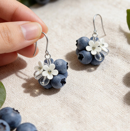 Blueberry Bouquet Earrings photographed with fresh blueberries and green leaves in soft natural light, with one earring gently held by hand for scale. Handmade in Finland by byMinta.