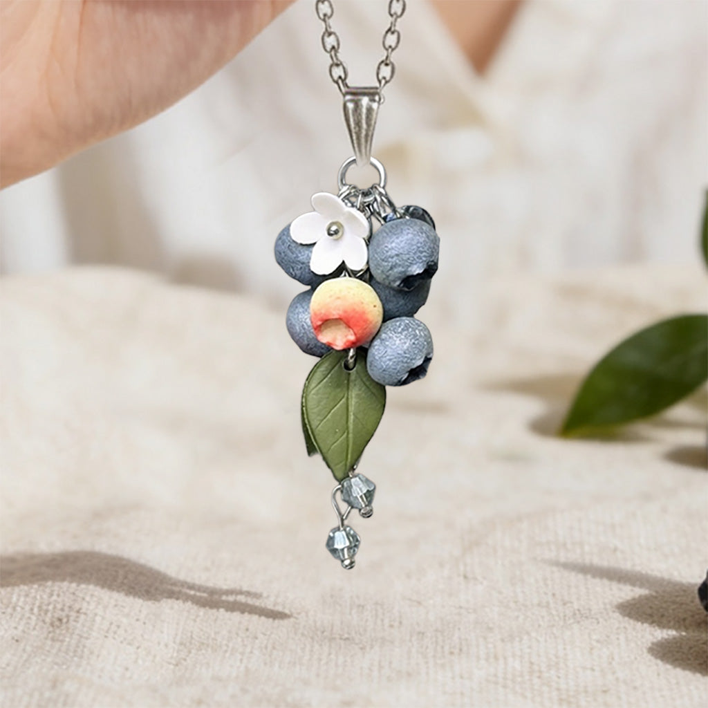Handmade berry necklace featuring sculpted blueberries in different stages of ripeness, translucent crystal beads, a green leaf and delicate white flowers. Botanical statement jewelry handcrafted in Finland.