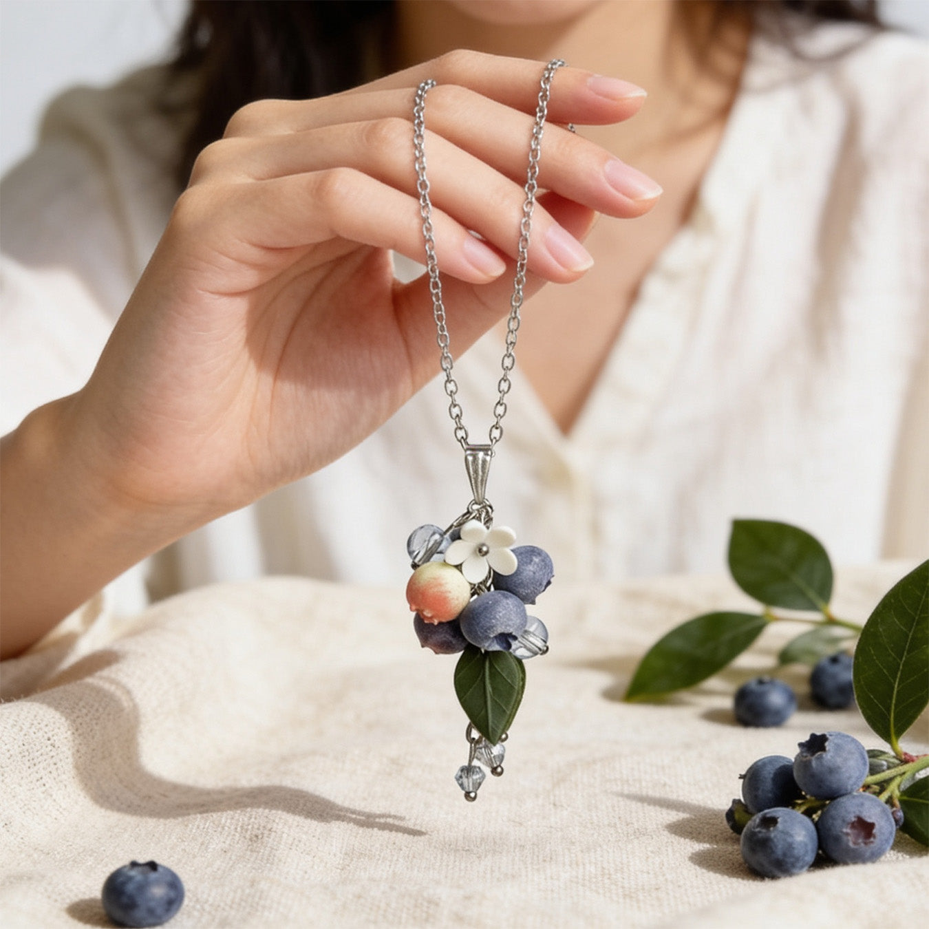Blueberry Branch Necklace held gently in hand for scale, featuring hand-shaped blueberries and leaves made from polymer clay. Natural linen fabric, fresh blueberries, and green blueberry branches in soft natural light. Handmade in Finland by byMinta.