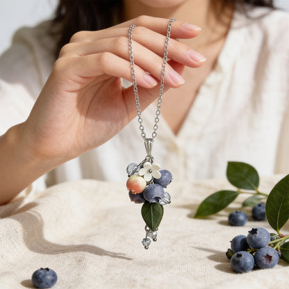 Blueberry Branch Necklace held gently in hand for scale, featuring hand-shaped blueberries and leaves made from polymer clay. Natural linen fabric, fresh blueberries, and green blueberry branches in soft natural light. Handmade in Finland by byMinta.