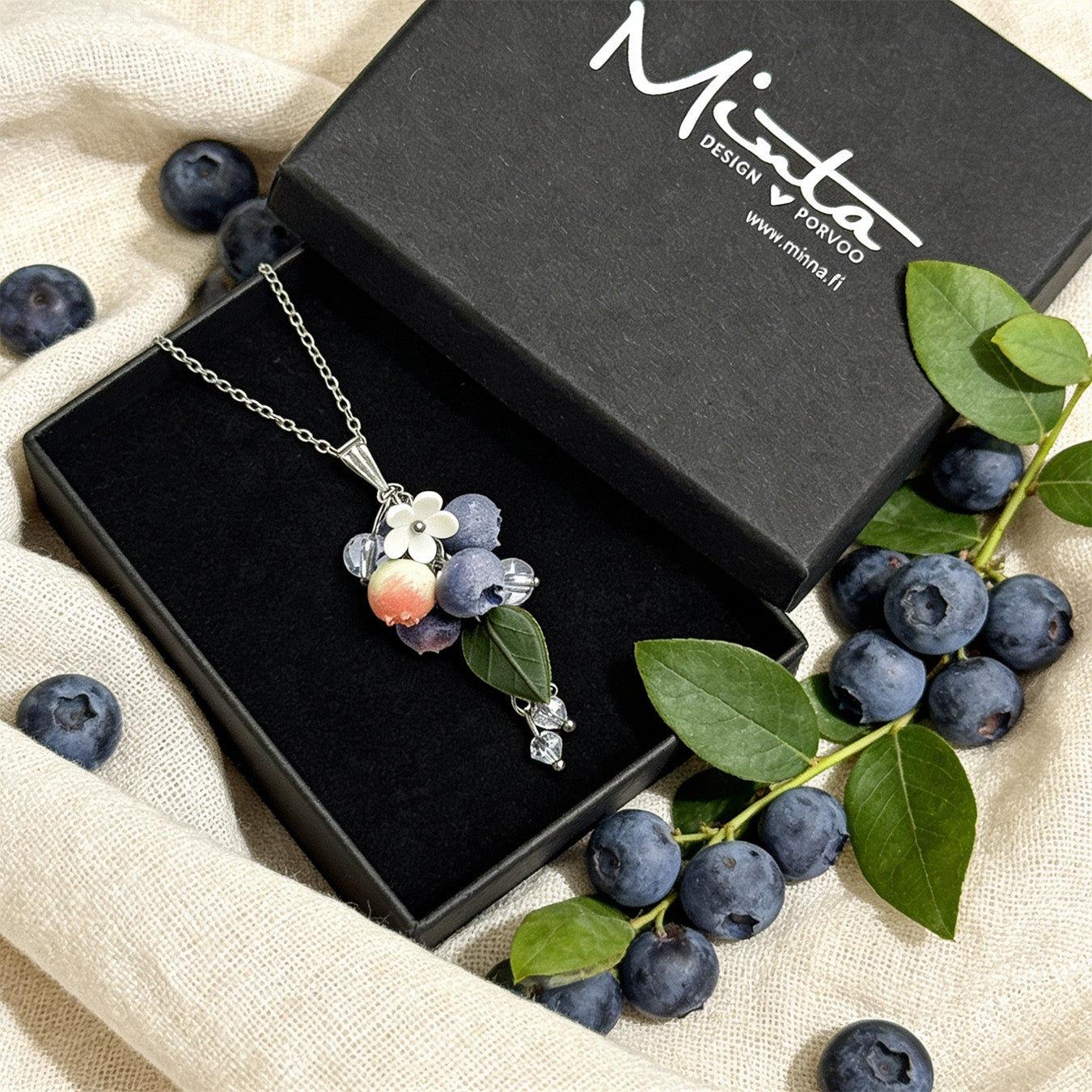 Blueberry Branch Necklace presented in a gift box, surrounded by natural linen fabric, fresh blueberries, and green blueberry branches in soft natural light. Handmade necklace with hand-shaped berries and leaves, handmade in Finland by byMinta.