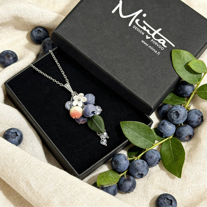Blueberry Branch Necklace presented in a gift box, surrounded by natural linen fabric, fresh blueberries, and green blueberry branches in soft natural light. Handmade necklace with hand-shaped berries and leaves, handmade in Finland by byMinta.