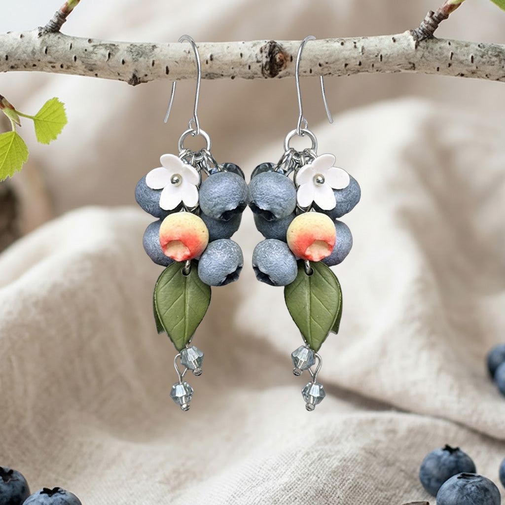 Handcrafted blueberry earrings hanging from a birch branch with white flowers and green leaves.