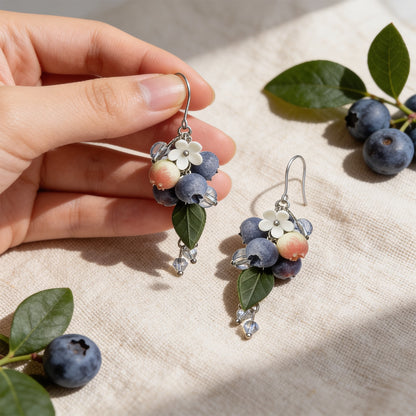 Blueberry Branch Earrings photographed with fresh blueberries and green leaves in soft natural light, with one earring gently held by hand for scale. Handmade in Finland by byMinta.