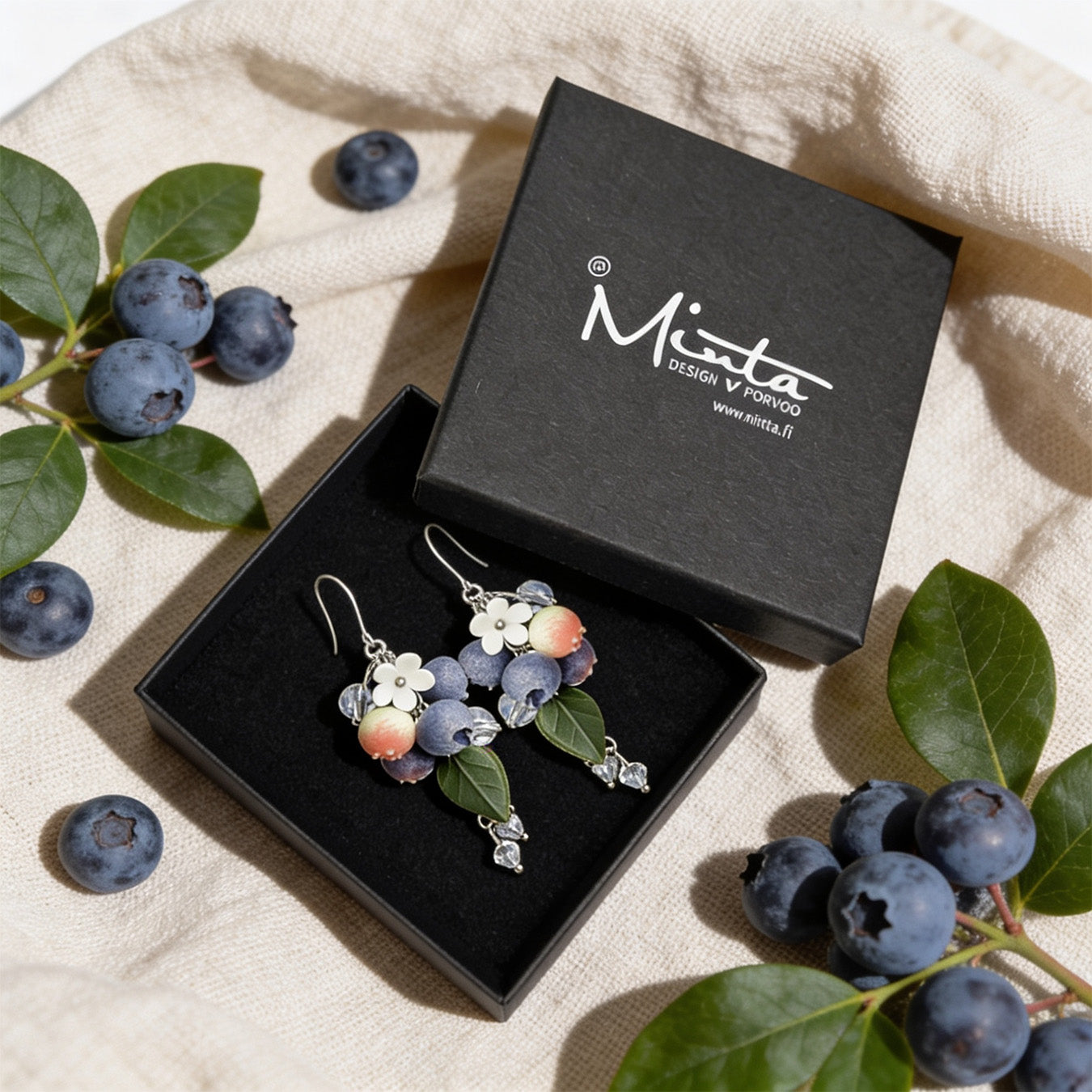 Blueberry Branch Earrings presented in a gift box, surrounded by natural linen fabric, fresh blueberries, and blueberry leaves. Handmade in Finland by byMinta.