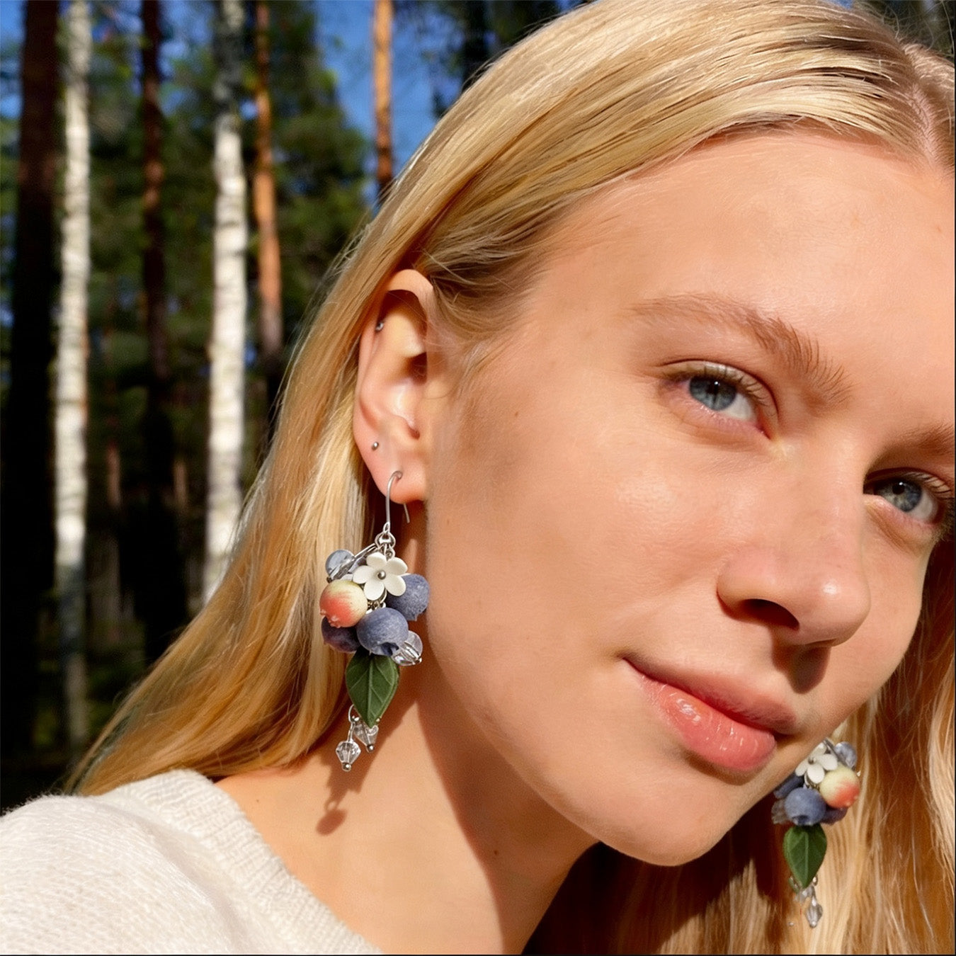 Blonde woman wearing Blueberry Branch Earrings in a sunlit Nordic forest. Handmade earrings inspired by the peaceful beauty of summer blueberries.