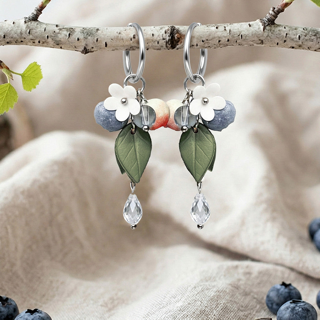 Berry Huggie hoop earrings with blueberries, flowers, leaves and crystal drops, handmade in Finland, photographed on a birch branch in a Scandinavian setting