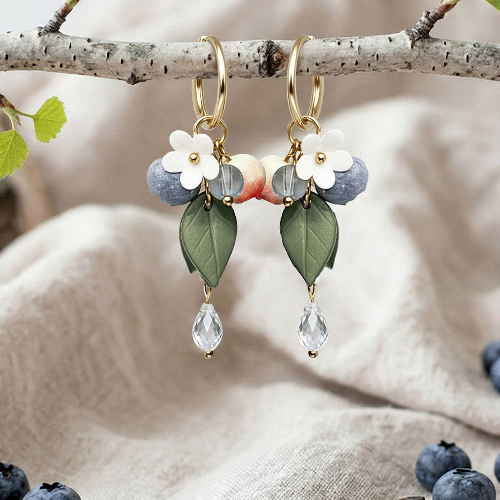 Berry Huggie hoop earrings with blueberries, flowers, leaves and crystal drops, handmade in Finland, photographed on a birch branch in a Scandinavian setting