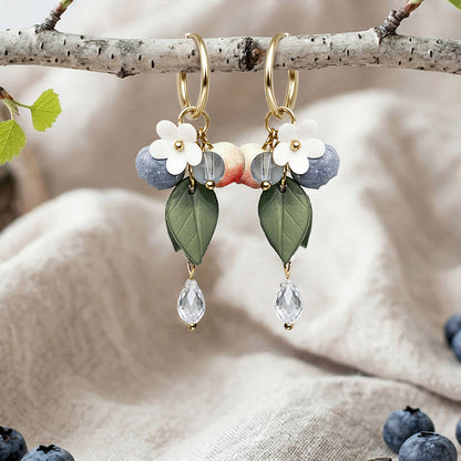 Berry Huggie hoop earrings with blueberries, flowers, leaves and crystal drops, handmade in Finland, photographed on a birch branch in a Scandinavian setting