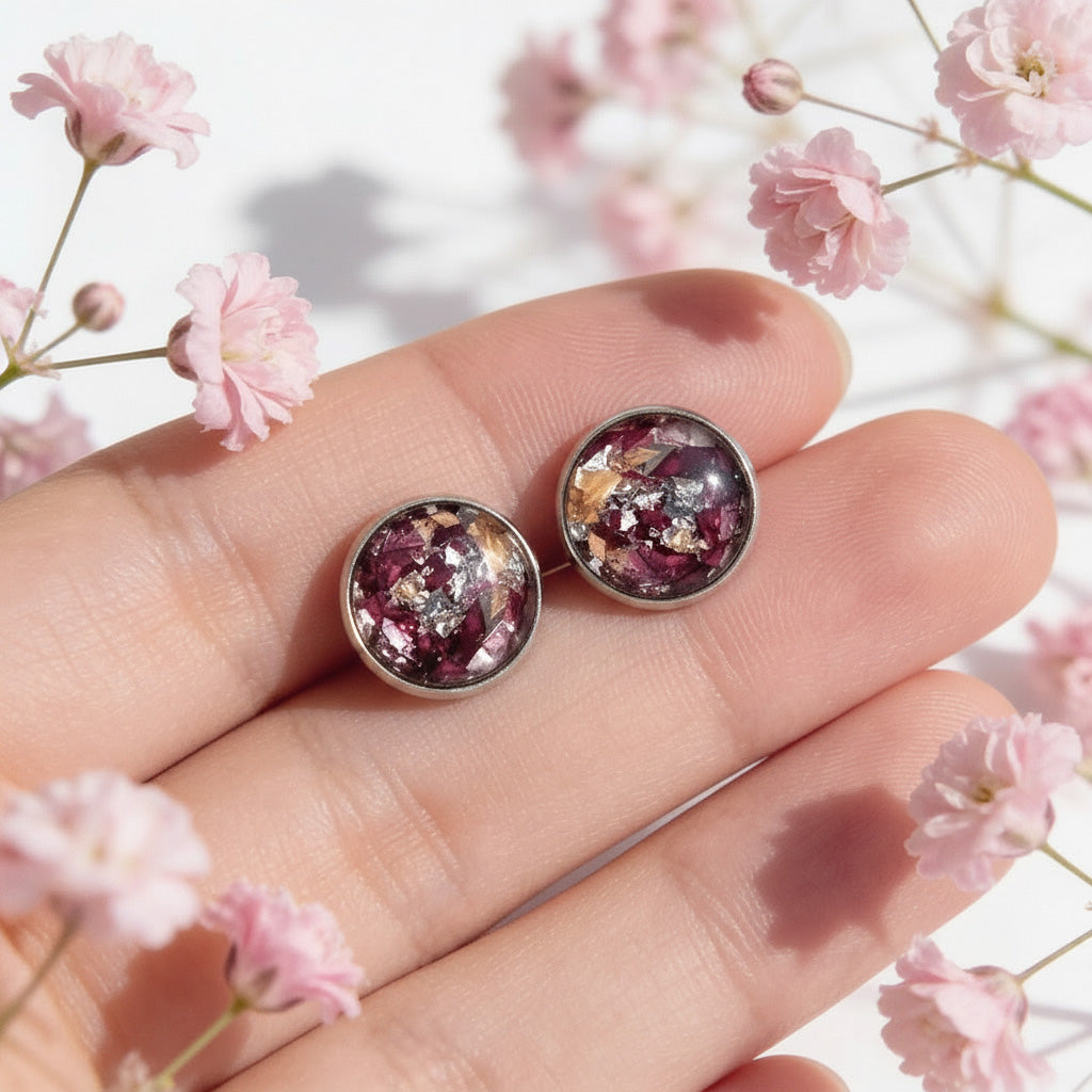 Handmade rose petal stud earrings with real preserved rose petals and silver leaf, photographed on fingertips with soft pink flowers in natural light.