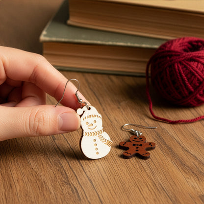 Hand holding the snowman earring on a wooden table with vintage books and burgundy yarn, with the gingerbread earring beside it. Handmade in Finland by byMinta.