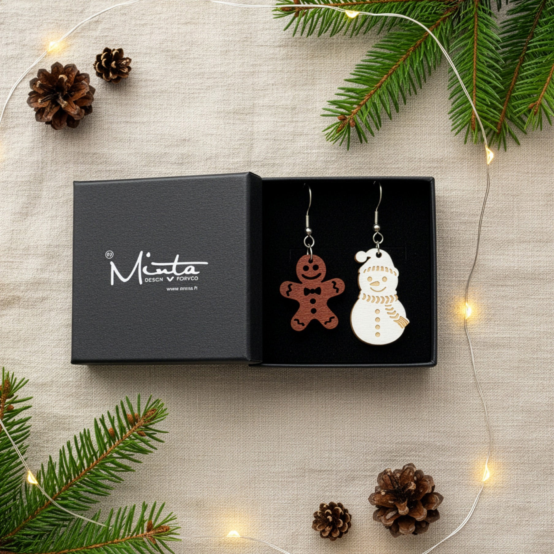 Gingerbread and snowman earrings presented in a gift box with pine branches, pinecones, and warm fairy lights, creating a cozy festive atmosphere. Handmade in Finland by byMinta.