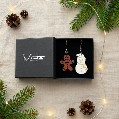 Gingerbread and snowman earrings presented in a gift box with pine branches, pinecones, and warm fairy lights, creating a cozy festive atmosphere. Handmade in Finland by byMinta.