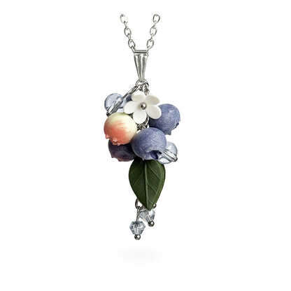 Handmade berry necklace featuring sculpted blueberries in different stages of ripeness, translucent crystal beads, a green leaf and delicate white flowers. Botanical statement jewelry on a fine chain against a clean white background, handcrafted in Finland.
