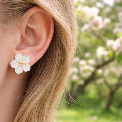 Woman wearing white apple blossom stud earrings by byMinta in soft natural daylight with blooming spring flowers in the background.