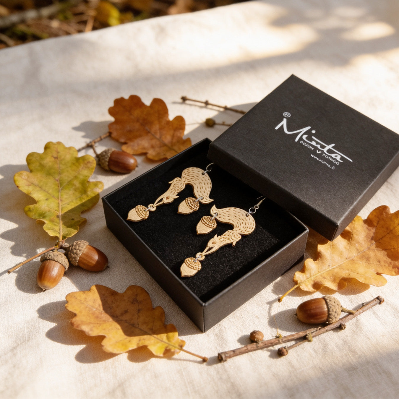 Handmade squirrel earrings displayed in a black jewelry box surrounded by oak leaves, acorns and small twigs. Lightweight Finnish birch plywood earrings styled in warm natural daylight with a soft Nordic autumn forest atmosphere.