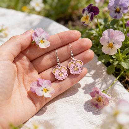 Hand holding handmade mauve pansy earrings for size reference among real pansy flowers. Lightweight Finnish birch plywood floral earrings photographed in soft natural sunlight with a fresh Nordic spring garden atmosphere.