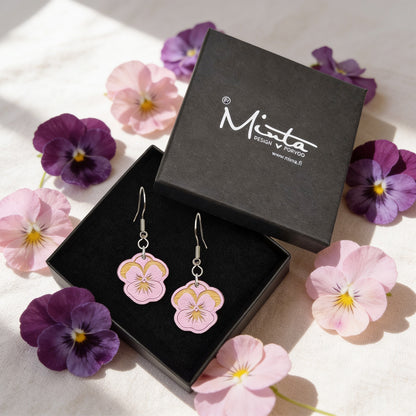 Handmade mauve pansy earrings displayed in a black jewelry box surrounded by real pansy flowers. Lightweight Finnish birch plywood floral earrings styled in soft natural daylight with a fresh Nordic spring atmosphere.