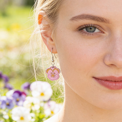 Close-up portrait of a Scandinavian woman wearing handmade mauve pansy earrings in a blooming spring garden. Lightweight Finnish birch plywood floral earrings photographed in soft natural sunlight with a fresh Nordic atmosphere.