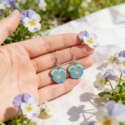 Hand holding handmade sky blue pansy earrings for size reference among blooming pansy flowers. Lightweight Finnish birch plywood floral earrings photographed in soft natural sunlight with a fresh Nordic spring garden atmosphere.