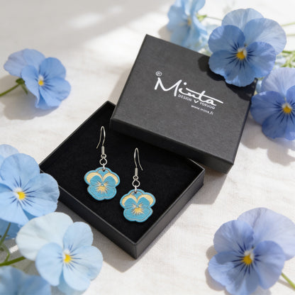 Handmade sky blue pansy earrings displayed in a black jewelry box surrounded by real blue pansy flowers. Lightweight Finnish birch plywood floral earrings styled in soft natural daylight with a fresh Nordic spring atmosphere.