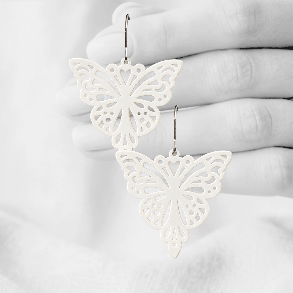White Papillon silicone earrings made in Finland, photographed against a black and white background of a hand.