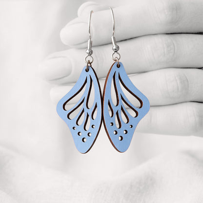 Light Blue Perhosensiipi butterfly wing earrings made from Finnish birch plywood, photographed hanging from a hand against a softly blurred light background.