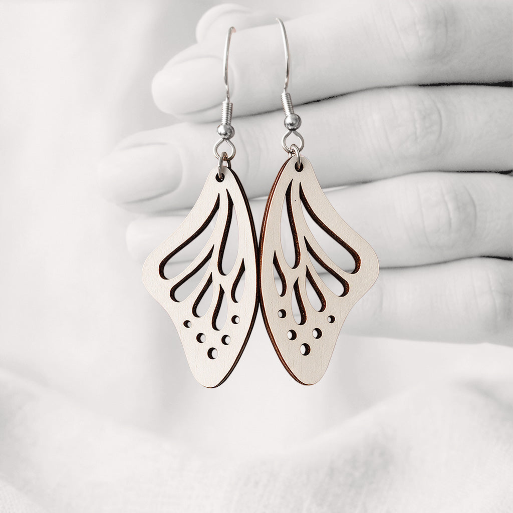 White Perhosensiipi butterfly wing earrings made from Finnish birch plywood, photographed hanging from a hand against a softly blurred light background.