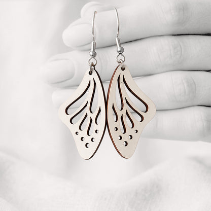 White Perhosensiipi butterfly wing earrings made from Finnish birch plywood, photographed hanging from a hand against a softly blurred light background.