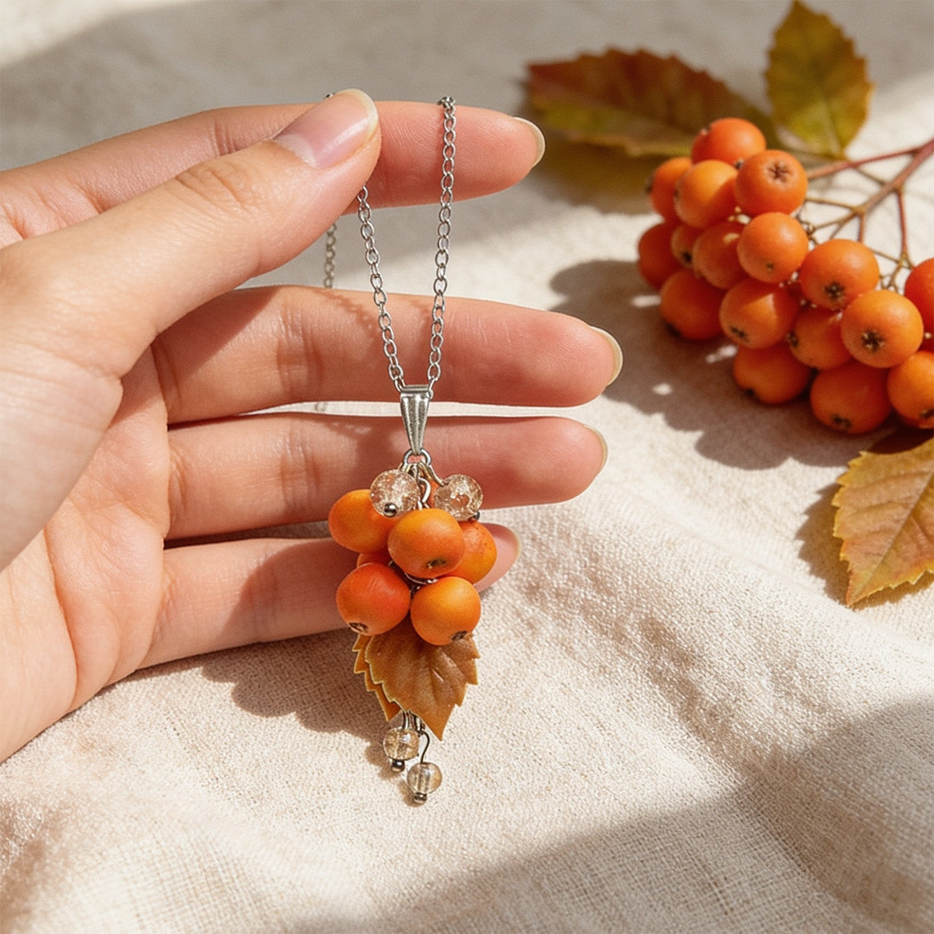 Rowan Berry Necklace held gently in hand for scale, featuring hand-shaped orange berries and leaves. Natural linen fabric, rowan berries, and autumn leaves in soft natural light. Handmade in Finland by byMinta.